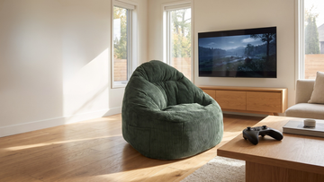Grand pouf vert émeraude en velours côtelé dans un salon avec une manette de jeu sur une table basse devant et une télé allumé avec un jeu.